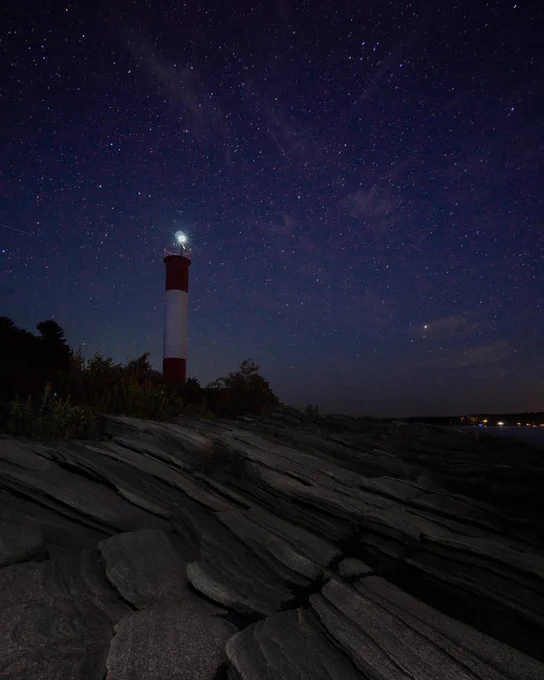 A nighttime photo of a lighthouse with a red and white striped tower under a starry sky, standing on rocky terrain.