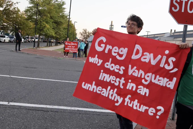 A couple people stand in the foreground, holding a banner that says "Greg Davis, why does Vanguard invest in an unhealthy future?" while more people with another banner stand across the street. Photo by Laran Kaplan