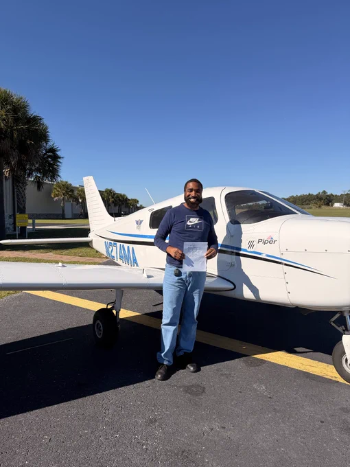 A black man wearing a blue Nike shirt and blue jeans, standing in front of a small piper airplane holding a private pilot certificate