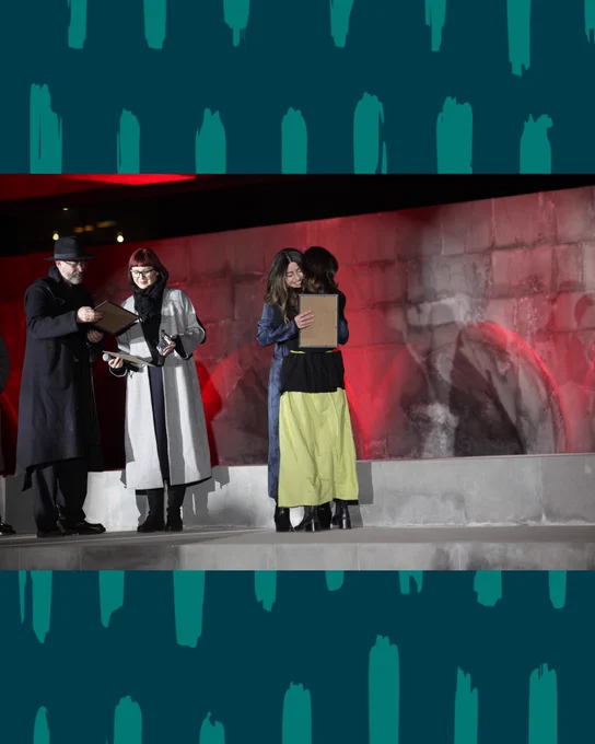 Two women embrace warmly on a stage while holding certificates, with a man and another woman standing nearby, all dressed in winter coats against a stone wall lit with red light.