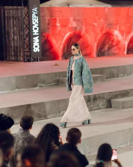 Model walks down concrete steps wearing a teal oversized jacket and white flowing skirt with matching platform shoes at a fashion event.