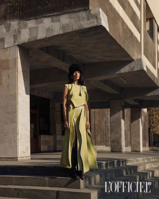 Model wearing a stylish olive green sleeveless dress with side slits and black boots standing on concrete steps under a modern building's overhang.
