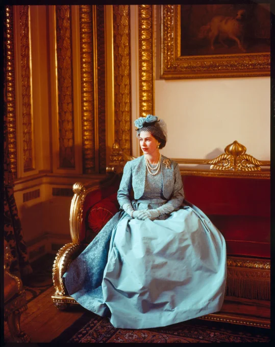 Queen Elizabeth II sitting on an ornate chair in a richly decorated room, wearing a light blue gown and matching hat.