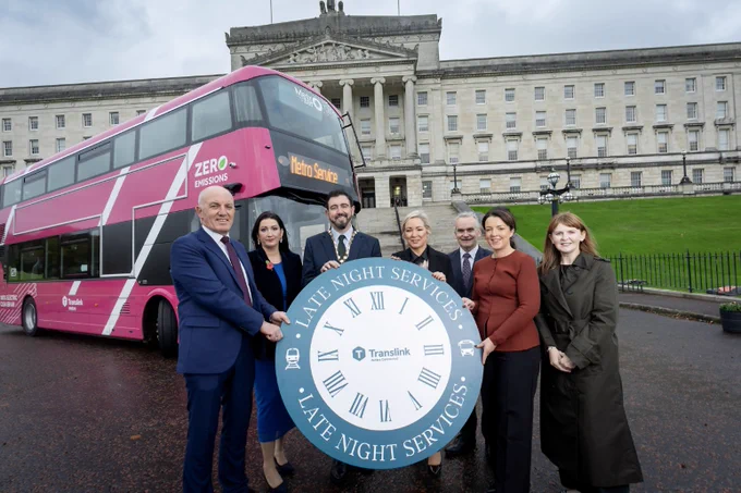 Image taken at Stormont, with Metro bus in the background & local political  & Translink representatives holding a large clock face with the Translink logo in the centre & late night services written around the outside. 
