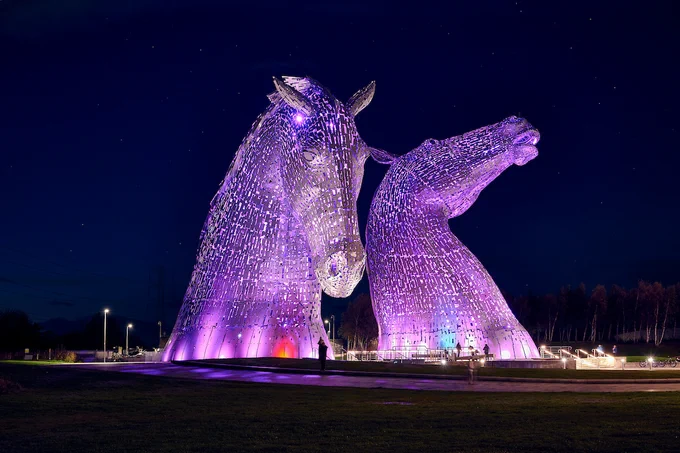 Two giant metal horse sculptures known as 'the Kelpies' illuminated in purple for World Stroke Day against a black sky.   
