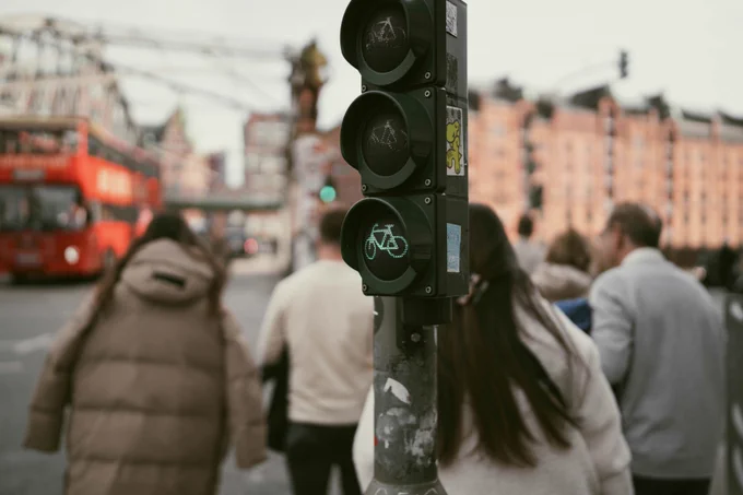A close up of a traffic signal with green cycle light illuminated. Blurred background image of people walking, buildings and a red bus.