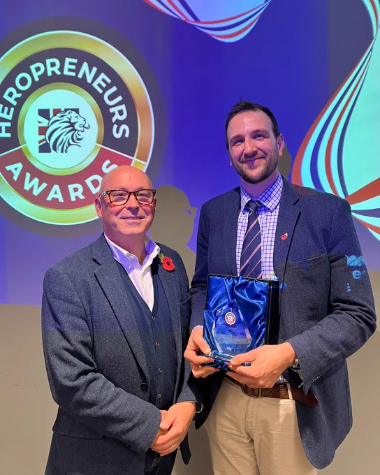 Two men in suits stand in front of a Heropreneurs Awards backdrop, one holding a glass award wrapped in blue fabric.