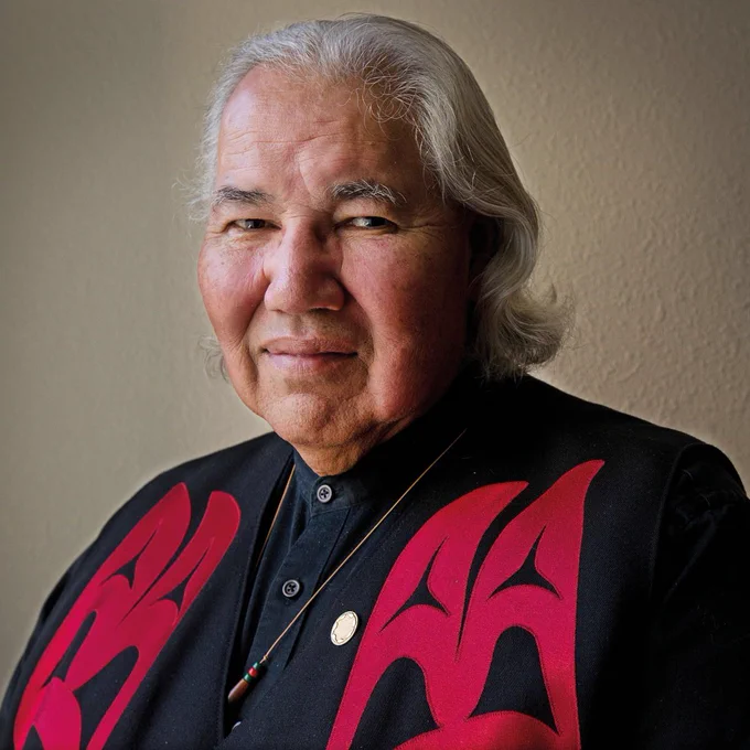 A person with long, gray hair wearing a black shirt with bold red designs, a necklace with a round pendant, and a beaded ornament, standing against a plain background.