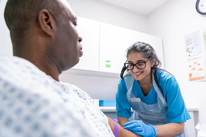 A healthcare professional in blue scrubs smiles while preparing to take a blood sample from a patient wearing a hospital gown in a clinical setting.