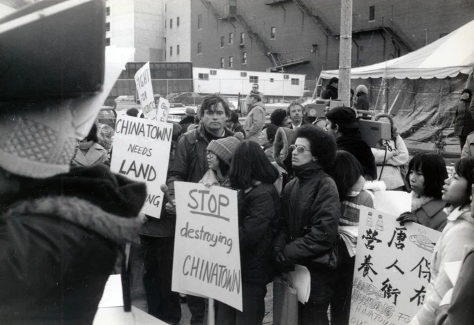 Black and white image of a group of protestors standing in a street. They are holding signs in English and Chinese, with English signs reading "Stop destroying Chinatown" and "Chinatown needs land."