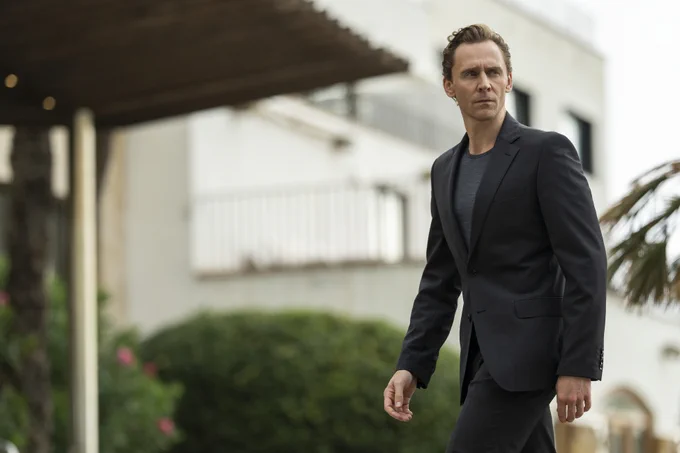 A man in a dark suit walks outdoors looking seriously toward something out of shot