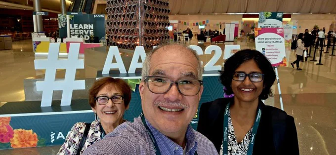 Three individuals smiling at the camera in a conference hall with a hashtag sign "AAMC25" prominently displayed in the foreground.