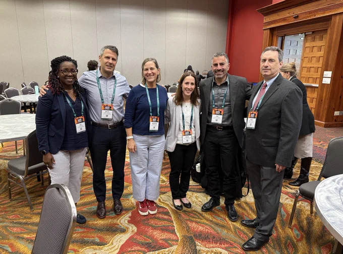 Group of six professionals smiling at a conference, wearing badges and casual to business attire.