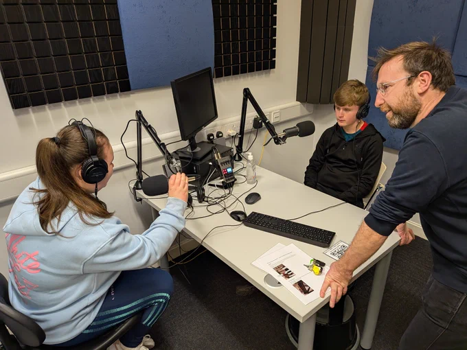 Photo of two secondary school students getting a guided tour of UCC Boole Lib podcast studio from Declan Synnot with the student wearing headphones and exploring the mics