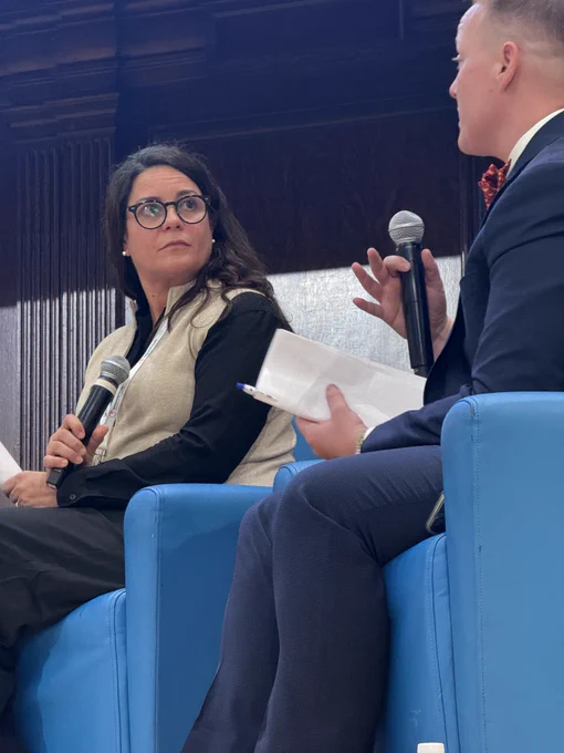 Josiane Martinez, a Latina woman, answers a question from Alexandria Eberhardt on stage. Alexandria is holding a microphone and is on the right side. Josiane is wearing dark rimmed glasses and looking at Alexandria from the left side.
