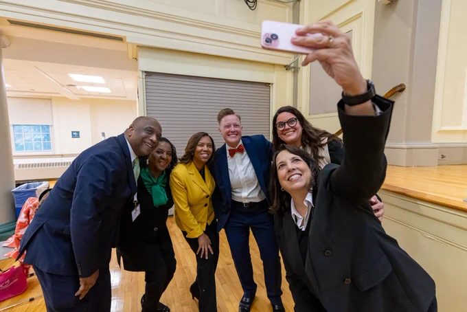 Six people group together to take a selfie. On the far left is is a Black man with close-cropped hair wearing a suit and green tie; to his right is a Black woman with short hair wearing a green scarf; to her right is Sabina Matos, wearing a yellow blazer; to her right is Alexandria Eberhardt, wearing a blue suit and red bowtie; to her right is Josiane Martinez; and in front is Kim Driscoll holding a phone and taking a selfie with all of them.
