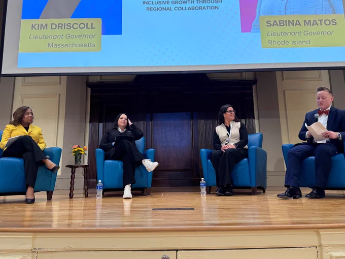 Four people sitting on blue chairs and presenting under a screen that reads "Kim Driscoll, Lieutenant Governor, Massachusetts. Inclusive Growth Through Region Collaboration. Sabina Matos, Lieutenant Governor, Rhode Island . On the far left is Sabina Matos, a Black woman with shoulder-length hair wearing a yellow blazer and black slacks. Second from left is Kim Driscoll, a white woman wearing a Black blazer and black pants. Third from left is Josiane Martinez, a Latina woman wearing a tan vest and black pants. On the far right is Alexandria Eberhardt, a white woman wearing a blue suit and red bowtie.