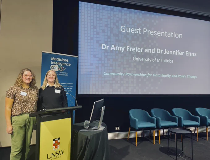 Photograph of two people standing behind a podium with a yellow banner with a crest and the letters UNSW visible. One has curly, shoulder length blonde hair and is wearing a patterned top, beside is a person with long, straight blonde hair, wearing a black top. Both have visible nametags. Beside them a video screen with a row of chairs in front displays the words Guest Presentation, Dr. Amy Freier and Dr. Jennifer Enns, University of Manitoba.