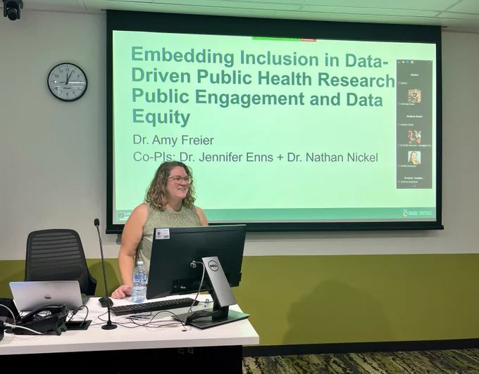Image of a person with curly blonde hair and glasses, standing behind a desk with a computer monitor facing them. Behind is a video screen showing the words "Embedding Inclusion in Data-Driven Public Health Research, Public Engagement and Data Equity, Dr. Amy Freier, Co-PIs, Dr. Jennifer Enns   Dr. Nathan Nickel.