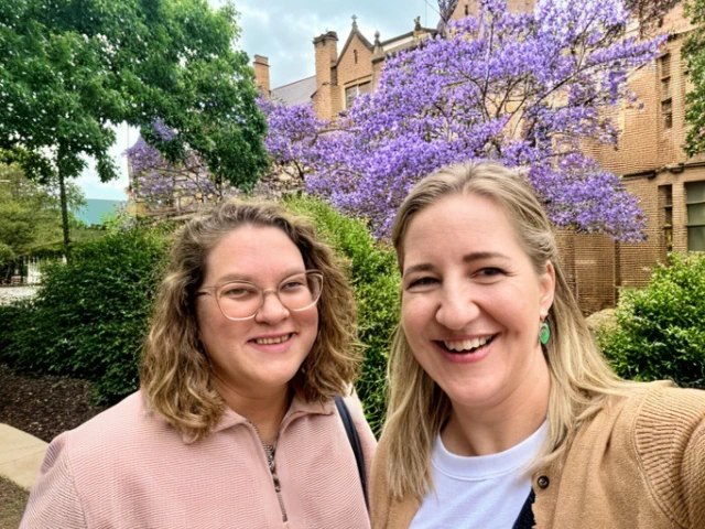 Image of two smiling people, outdoors, in front of green foliage, with a large tree covered in purple flowers behind, and a gothic-style building further back. One person has long, straight blonde hair, a beige cardigan and green earrings, the other has wavy, shoulder length blonde hair and glasses, wearing a dusty pink pullover top.