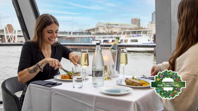 Two women sat at a table with a white table cloth, drinks and food with the River Thames view in the background.