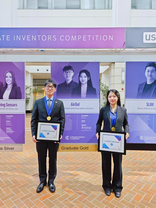 Two people dressed in black suits stand on a light brown brick floor holding framed awards with blue and purple designs. They wear gold medals with blue ribbons around their necks. Behind them is a large purple and white display that reads “Collegiate Inventors Competition” and features their project “AirGel,” shown with a gradient of purple and blue tones. 

Image Courtesy of Weixin Guan.