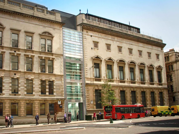 View of the exterior of a building in London with pedestrians walking by and a red double-decker bus on the street.