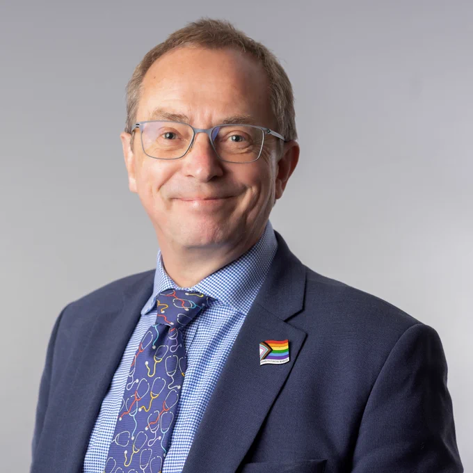 Dr Jim Gardner,  Group Chief Medical Officer at NHS University Hospital of Liverpool Group, wearing a blue suit jacket, patterned tie, and a rainbow flag pin on his lapel. Jim is smiling whilst standing against a plain grey background.