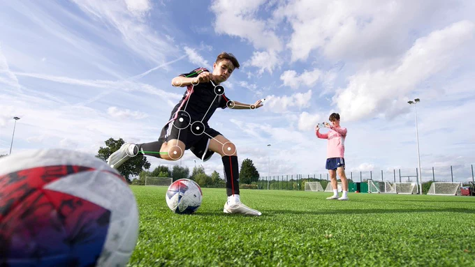 A person on a football pitch is captured mid-kick, wearing a black sports outfit and white shoes, against a clear blue sky with scattered clouds overhead. Motion-tracking markers are visible on the body's joints. Another person in a pink top and dark shorts stands in the background, holding a phone. 