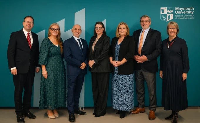 A group of seven people, four women and three men, stand smiling in front of a teal backdrop with the Maynooth University logo. They are dressed smartly in formal attire, with a mix of suits and dresses.