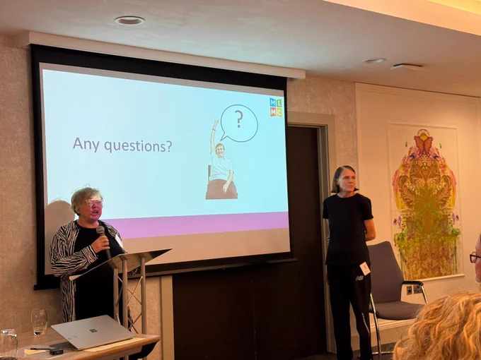 A photo of Simone Aspis standing in front of a projector screen, which shows the words "Any questions?" with a photo of a person raising their hand. Another woman is standing to her right. 