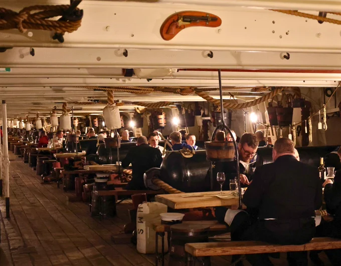 People dining under the deck of the HMS Warrior, with tables set amongst historical naval artifacts and ambient lighting.
