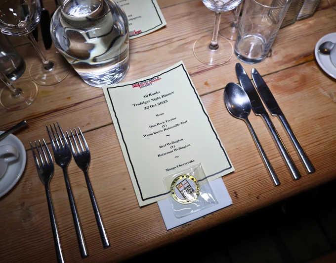 A formal dining setting featuring a neatly arranged silverware set and a menu titled "Royal Navy & Royal Marines Charity" on a wooden table. Next to the menu is a coin with the RN emblem. 
