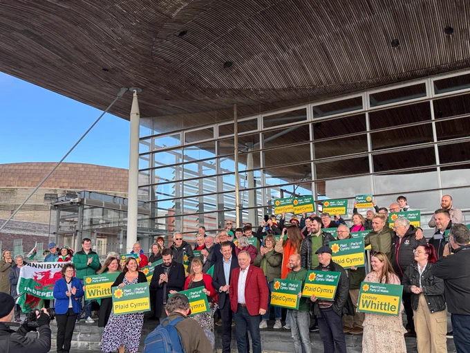 A large crowd of Plaid Cymru MSs and activists holding placards in front of the Senedd huilding.  Lindsay Whittle is stood in the front at the centre.