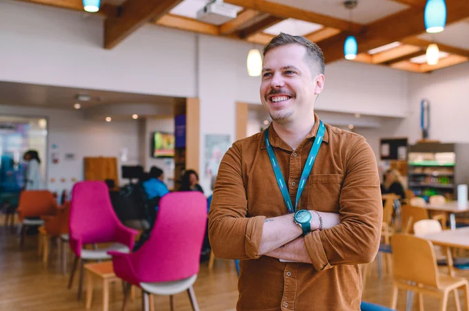 Person smiling and standing with arms crossed in a busy cafeteria setting.