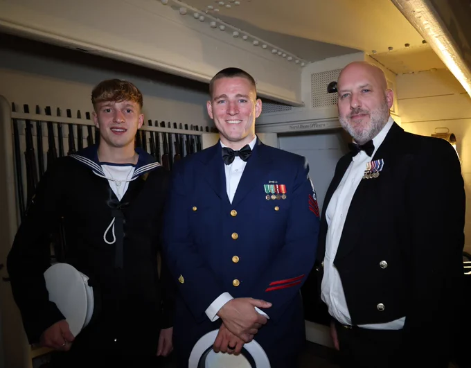 Three individuals in formal military attire smiling at a social event. From left to right: a person in a navy sailor uniform, a person in a Coast Guard dress uniform with medals, and a person in a dark suit with medals.