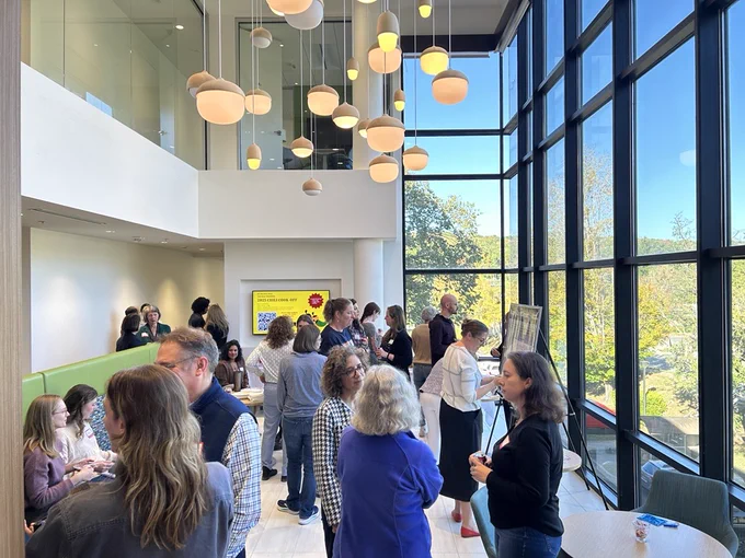 People mingling in a room with large windows and large lights hanging down from the ceiling.