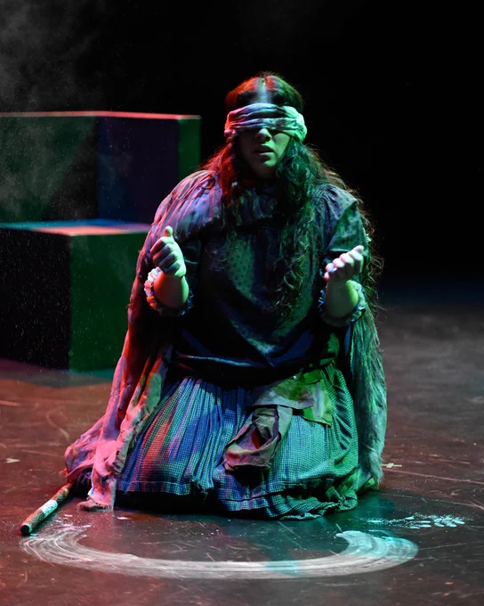Performer in a colorful costume sitting on stage with a painted circle, blindfolded, and holding a stick during a theatrical production.