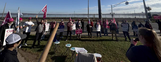 Many union and faculty associations gathered by the ocean listening to a speaker with a microphone. Most are carrying signs or flags.