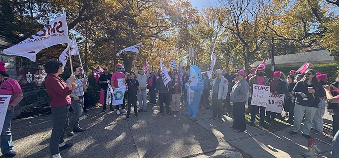 A large gathering of union and faculty association members carrying flags and placards with the morning sun beaming in through the tree canopy.