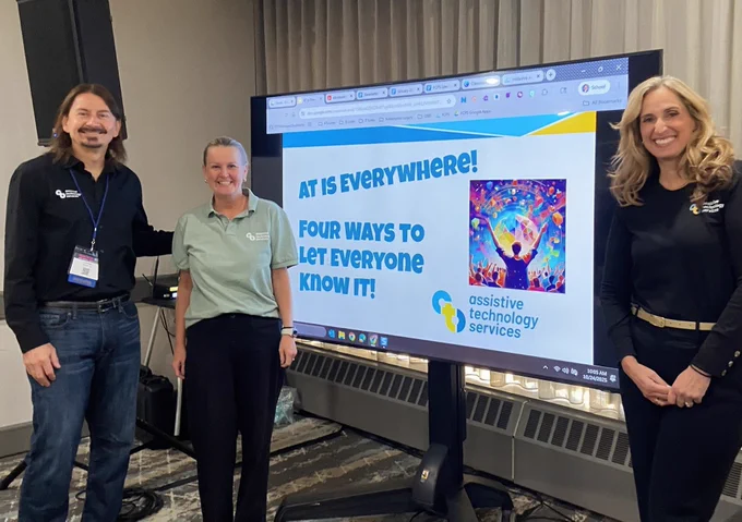 A man and a woman are standing to the left of a presentation board while an additional woman is standing to the right. They are all smiling. The scree has text stating "AT Is Everywhere! Four Ways to Let Everyone Know It!"