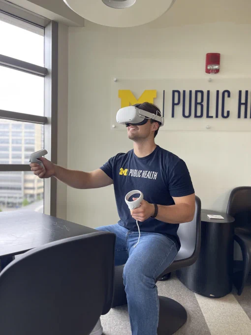 Person in a 'Michigan Public Health' T-shirt using a virtual reality headset and controllers in a modern room with a large University of Michigan School of Public Health logo on the wall.