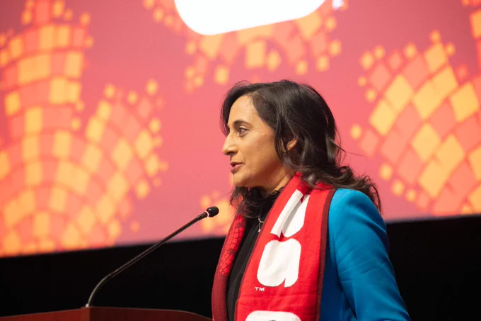Minister Anita Anand speaking at a podium, wearing a blue blazer and a red scarf with white lettering, with a patterned backdrop behind her.
