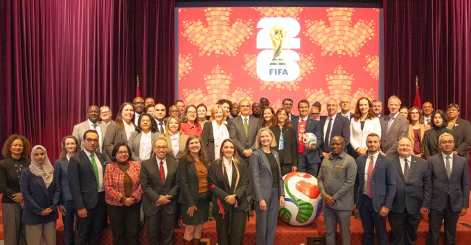 A large group of people standing on a stage in front of a red backdrop featuring the FIFA World Cup 2026 logo. Two oversized soccer balls are placed on the floor in front of the group.