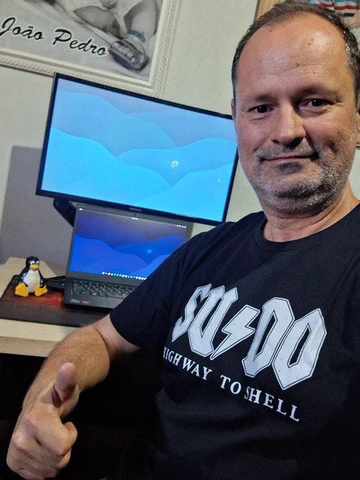 Um homem sorri fazendo sinal de positivo em frente a um setup com notebook e monitor exibindo o fundo azul do Debian. Ele veste uma camiseta preta com a frase “SUDO HIGHWAY TO SHELL”, em referência divertida ao comando sudo do Linux e à música do AC/DC “Highway to Hell”. Ao lado do notebook há um boneco do Tux, o pinguim mascote do Linux. Em outra imagem, a mesma camiseta está esticada sobre uma superfície floral, com o Tux apoiado sobre ela.
