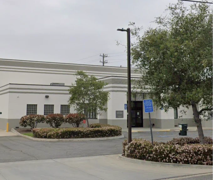 A white building with gray trim. There are flowering bushes and trees out front.