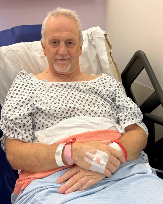 A hospital patient sitting in a bed and smiling at the camera, wearing a hospital gown.