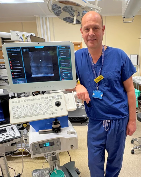 A healthcare professional in scrubs stands beside an ultrasound machine in a medical room.