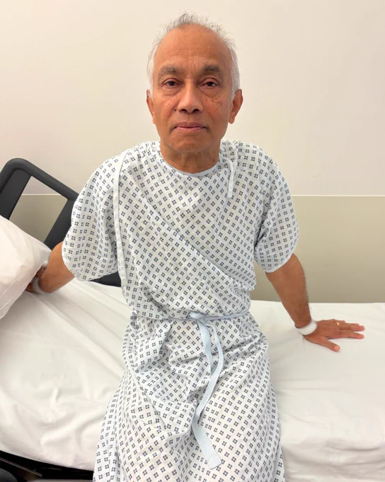 Patient sitting on a hospital bed wearing a standard hospital gown, looking directly at the camera.