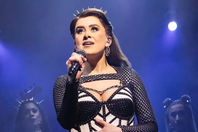 A photo showing a woman dressed as a trendy queen standing on a blue-lit stage, singing into a microphone 